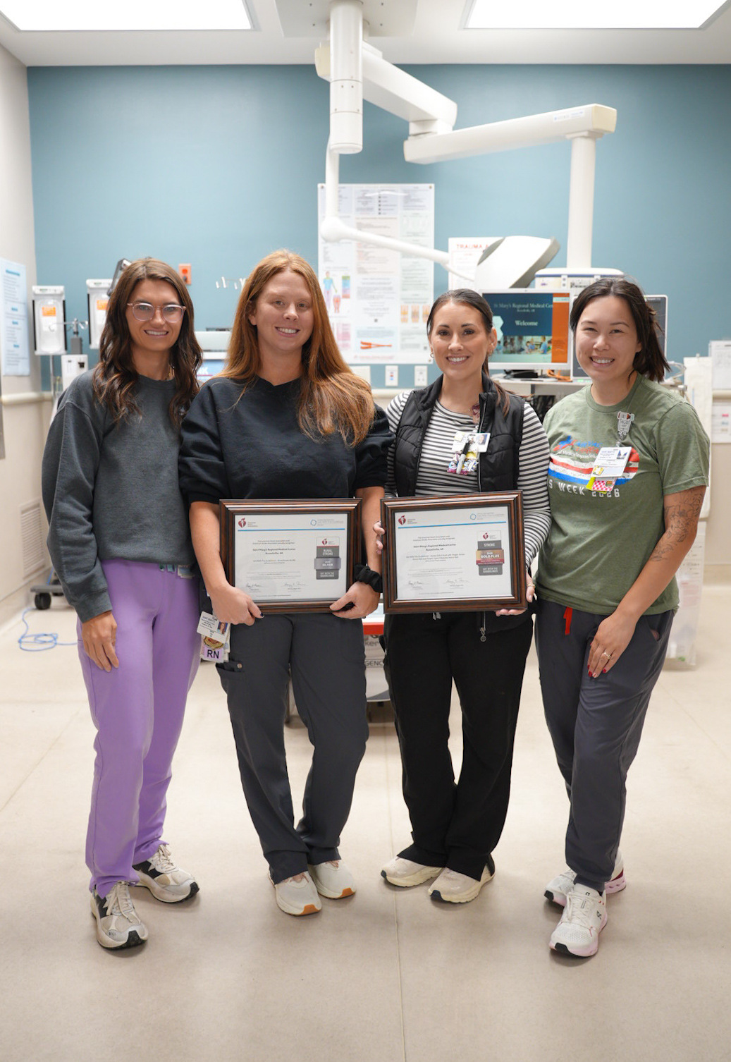 Four healthcare team members stand in a clinical treatment room holding framed American Heart Association stroke care awards. Medical equipment and monitors are visible in the background.