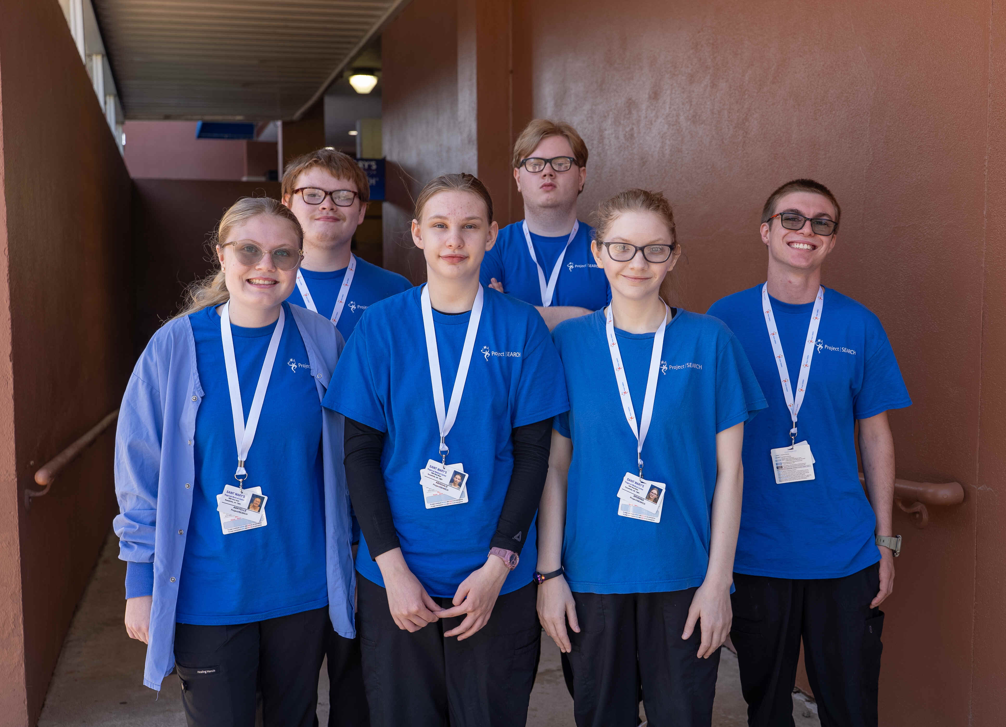 Six Project SEARCH interns wearing blue program shirts and hospital ID badges stand together in a covered outdoor corridor. Three interns are in the front row and three in the back, with brown walls and handrails framing the group as they face the camera.