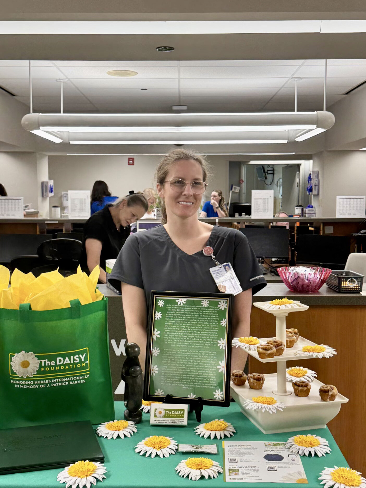 A Saint Mary’s nurse stands behind a DAISY Award display featuring a recognition plaque, daisies, treats and a DAISY Foundation gift bag at a nurses’ station. The nurse is being recognized for compassionate, exceptional patient care.