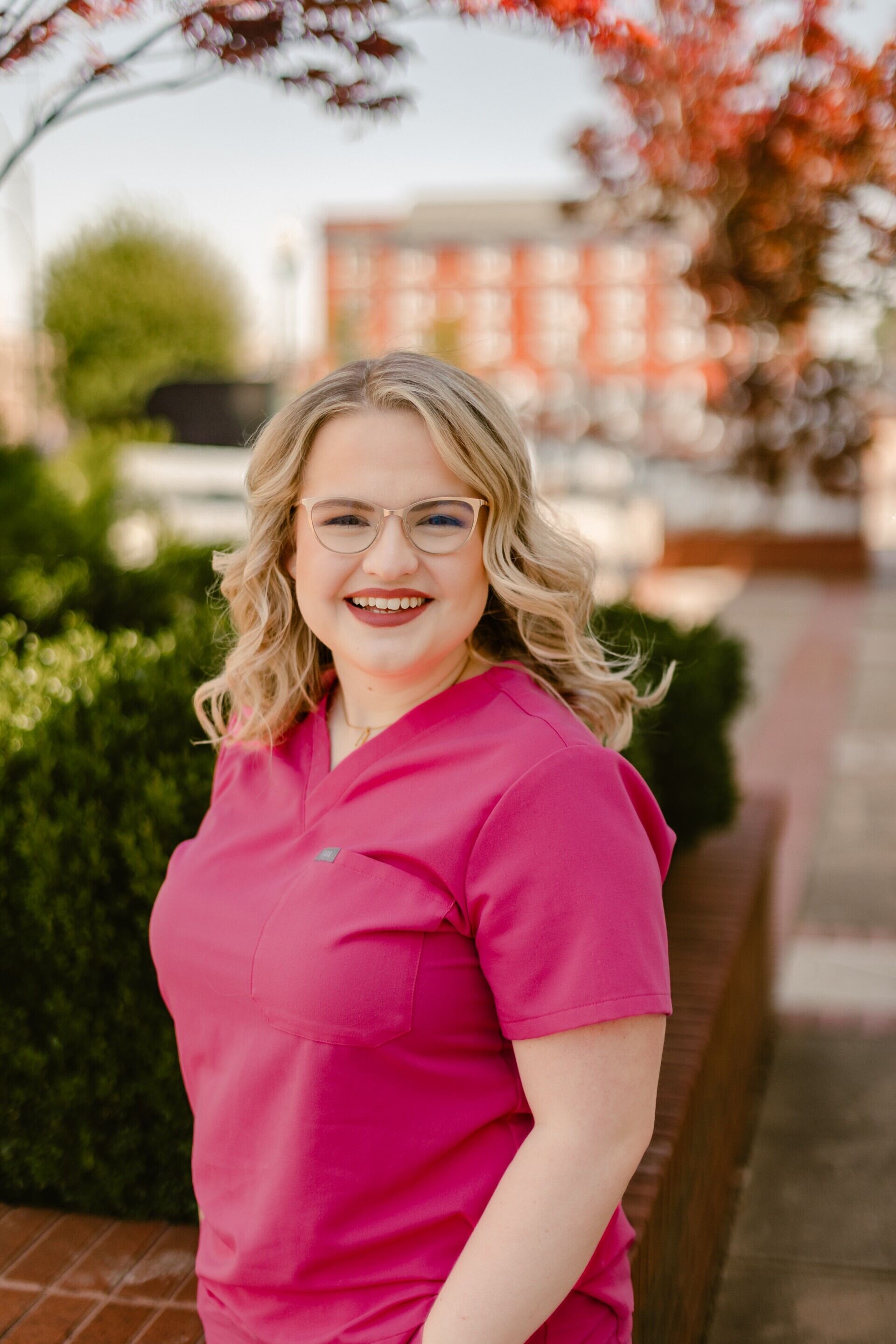 Professional outdoor headshot of a person in bright pink medical scrubs and glasses, standing beside a brick planter with greenery and a blurred building in the background.